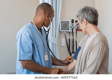 Male Nurse Checking Blood Pressure Of A Senior Woman In Medical Office. African Doctor Taking The Blood Pressure Of His Patient At Hospital. Young Doctor Measuring The Blood Pressure Of Mature Woman.