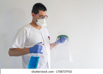 Male Nurse Cleaning A Face Shield. He Wears Gloves And A Mask. His Uniform Is White.