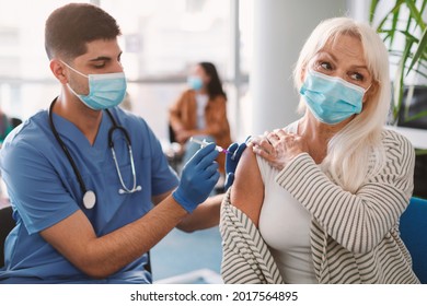 Male Nurse Or Doctor In Medical Face Mask Giving Injection To Older Female Patient During Mass Immunization Campaign At Health Center. Senior Woman Getting Covid Vaccine Shot At Clinic Or Hospital