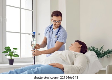 Male nurse gives an IV line to a female patient. Friendly young man in a scrubs uniform sets an intravenous infusions system for a happy overweight woman lying on the medical bed at the hospital - Powered by Shutterstock