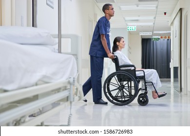 Male Nurse Pushing Patient Along Hospital Corridor In Wheelchair