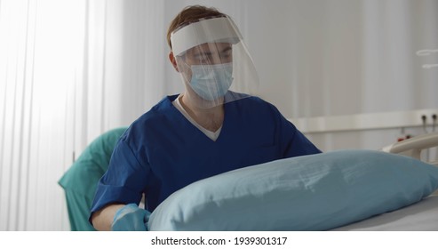 Male Nurse In Safety Gloves And Mask Working At Hospital Making Bed And Changing Sheets. Medical Staff In Safety Uniform Cleaning Empty Ward In Clinic During Coronavirus Pandemic