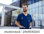 A male nurse is standing outdoors wearing scrubs and a stethoscope. He is smiling and looking at the camera. Portrait of male nurse. Portrait of mid adult nurse man at hospital