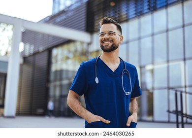 A male nurse is standing outdoors wearing scrubs and a stethoscope. He is smiling and looking at the camera. Portrait of male nurse. Portrait of mid adult nurse man at hospital