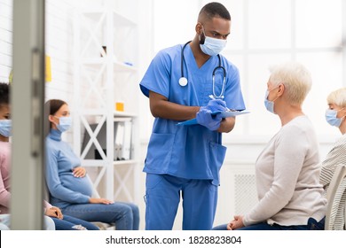 Male Nurse Talking With Senior Patient Lady About Covid-19 Symptoms Before Doctor's Appointment Sitting In Queue In Hospital, Wearing Face Masks. Coronavirus, Covid Medical Treatment Concept