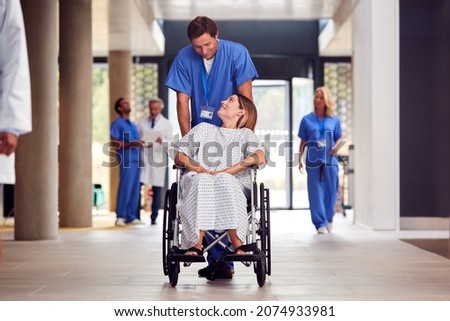 Male Nurse Wearing Scrubs Pushing Female Patient In Wheelchair Through Hospital Building Сток-фото © 