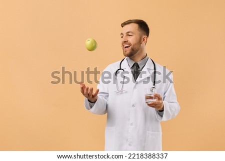 The male nutritionist doctor with stethoscope smiling and holding water and apple on beige background, diet plan concept