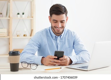 Male Office Worker Dressed In Blue Shirt Enjoying Break From Work, Scrolling News Feed On Phone, Having Fun At Content, Smiling Happily