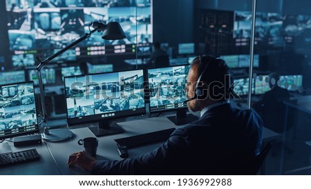 Male Officer Works on a Computer with Surveillance CCTV Video in a Harbour Monitoring Center with Multiple Cameras on a Big Digital Screen. Employees Sit in Front of Displays with Big Data.
