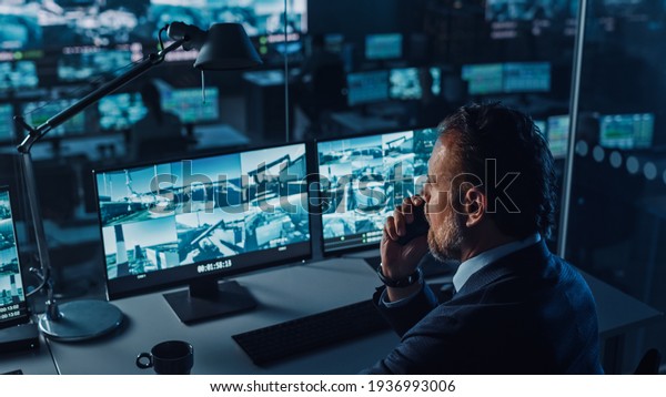 Male Officer Works on a Computer with\
Surveillance CCTV Video in a Harbour Monitoring Center with\
Multiple Cameras on a Big Digital Screen. Employee Uses Radio to\
Give an Order or Report.