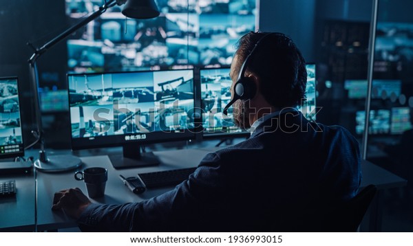 Male Officer Works on a Computer with Surveillance\
CCTV Video in a Harbour Monitoring Center with Multiple Cameras on\
a Big Digital Screen. Employees Sit in Front of Displays with Big\
Data.