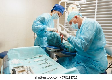 Male Old Professional Dentist Surgeon In Uniform And Female Assistant Helps To Performing Operation Install Dental Implant Teeth Of Woman Patient In Clinic Light Office With Modern Tools Equipment