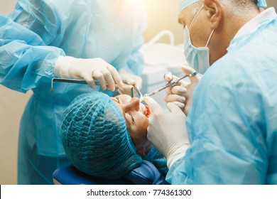 Male Old Professional Dentist Surgeon In Uniform And Female Assistant Helps To Performing Operation Install Dental Implant Teeth Of Woman Patient In Clinic Light Office With Modern Tools Equipment