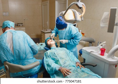 Male old professional dentist surgeon in uniform and female assistant helps to performing operation install dental implant teeth of woman patient in clinic light office with modern tools equipment