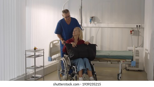 Male Orderly Pushing Senior Female Patient Being Discharged From Hospital In Wheelchair. Senior Man Nurse Pushing Wheelchair With Aged Woman Leaving Hospital After Recovery