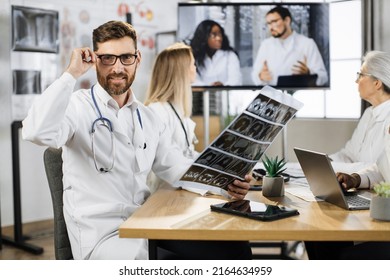 Male Orthopedist Holding Patient Result And Looking At Camera With Calm Face. Caucasian Medical Worker Having Distant Healthcare Seminar With Multiracial Coworkers In Clinic Meeting Room.