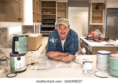 Male Painter Looking At Camera Holding Wet Paintbrush In Messy Home Kitchen, Fixer Upper Remodeling Project, Empty Paint Cans Stacked On Kitchen Counter, Lids Off