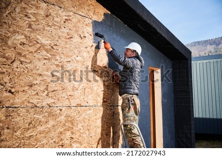 Male painter using paint roller, doing exterior paint work in a black color. Man worker building wooden frame house. Carpentry and construction concept.