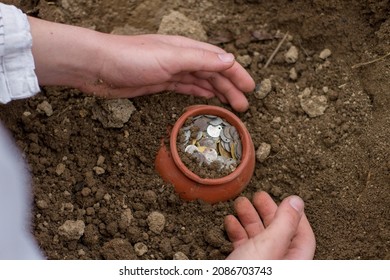 Male Pale Hand Digging Buried Treasure From The Dirt. Old Copper, Silver, And Gold Coins In A Pottery