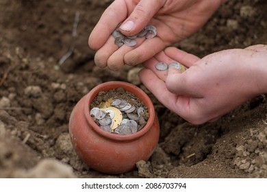 Male Pale Hand Digging Buried Treasure From The Dirt. Old Copper, Silver, And Gold Coins In A Pottery