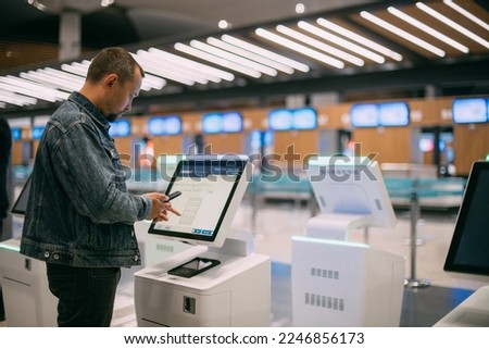 A male passenger at the electronic check-in desk in the departure area of the airport terminal.  A young guy checks in for a plane at the screen of a self-service computer kiosk to check in for flight