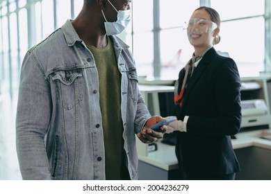 Male Passenger In Face Mask At Check-in Counter With Airport Staff. Airport Attendant Greeting Passenger After Check-in At Counter During Pandemic.