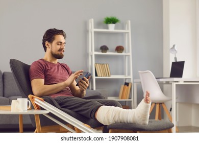 Male Patient With Broken Leg In Plaster Cast Relaxing At Home. Happy Man With Bone Fracture Sitting In Comfortable Armchair, Using Mobile, Texting Friends, Making Phone Calls, Chatting On Social Media