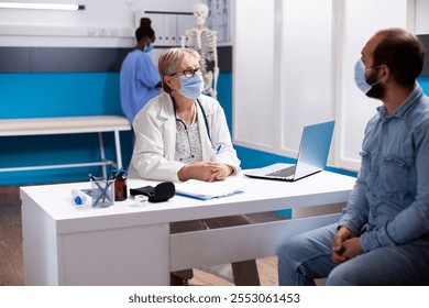 Male patient consults with senior doctor in hospital, both wearing face masks for safety. Laptop and clipboard placed on desk to support retired physician and young man during medical consultation. - Powered by Shutterstock