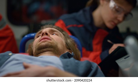 Male Patient Lying On Stretchers In Emergency Vehicle. Exhausted Man Feeling Pain In Stomach. Female Paramedic Writing Diagnosis On Clipboard. Ambulance Doctor In Uniform Working At Night Shift