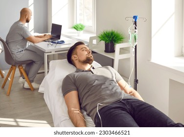 Male patient receiving intravenous therapy treatment at clinic. Young man getting vitamin or medication solution through IV line infusion while lying on medical bed in room with nurse in background