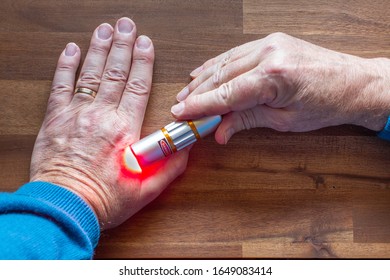 Male Patient Is Treating Himself On His Hand With Red Light From A Soft Laser For Health Application. A Warning Label Danger Is Attached To The Soft Laser Therapy Device.