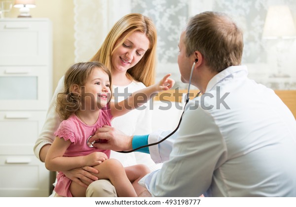 聴診器を持つ可愛い女の子を診察する男性小児科医 子どもは幸せそうにほほ笑み 先生を指差す 子供を抱いた母親 医者は自宅の患者を見舞う の写真素材 今すぐ編集