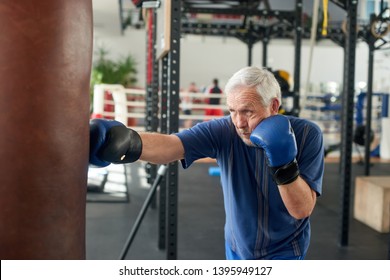 Male Pensioner Trains On A Punching Bag. Serious Older Man Working Out With Punching Bag At Boxing Hall. Sport Training Concept.