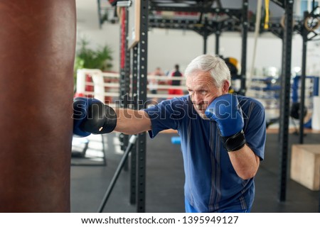 Male pensioner trains on a punching bag. Serious older man working out with punching bag at boxing hall. Sport training concept.