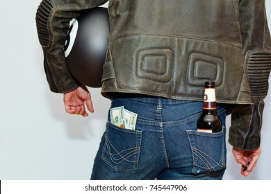 Male person in leather motorcycle protective jacket and blue jeans with motorcycle vintage style helmet with bottle of beer and stack of US dollars in pockets. Capture from back, no face.