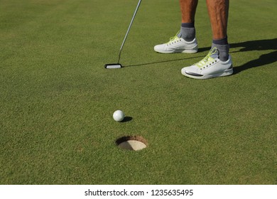 Male Person Playing Golf. Partial View Of The Legs, Sport Shoes, Socks And Bare Calves. Golf Club And A Golfball On The Green Grass. Moment After Putting The Ball On The Green, Rolling In The Hole.