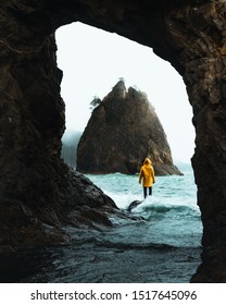 Male Person With A Yellow Rain Jacket Standing On A Rock At The Coast In Washington State. A Wave Is Coming In From The Sea Side And Causing Wet Clothes.