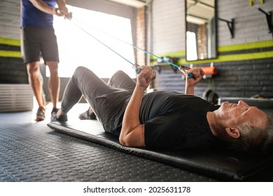 Male Personal Trainer Holds Onto Elastic Rubber Resistance Bands While An Elderly Senior Man Lying On Matt On Floor And Does Bicep Curl For Strength And Rehabilitation Exercise