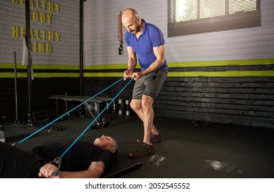 Male personal trainer holds onto elastic rubber resistance bands while an elderly mature man lying on exercise mat on floor and does bicep curl for strength and rehabilitation exercise with copy space