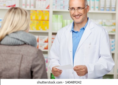 Male Pharmacist Holding Prescription Paper While Looking At Customer In Pharmacy