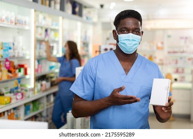 Male Pharmacist In Mask Holding Drug Package In Hands. His Colleague Working Behind.