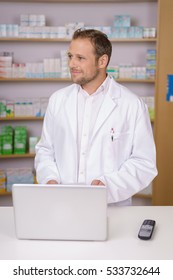 Male Pharmacist In A White Lab Coat Standing Behind A Counter With A Laptop Computer Watching Someone In The Pharmacy