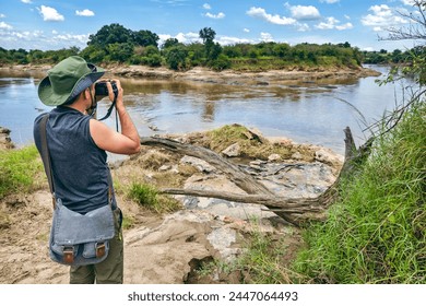 A male photographer in a green hat takes a photo the savannah on the background of river in masai mara, Kenya. The concept of an African walking safari in Africa. - Powered by Shutterstock