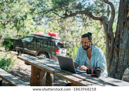 Male photographer working on a laptop outdoors in a camping. Guy freelancer, travel blogger working online at a laptop computer while hiking in the mountains outdoors. Work from anywhere in the world