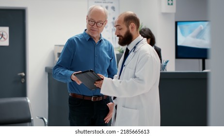 Male Physician Doing Consultation With Retired Man In Hospital Reception Lobby, Talking About Healthcare In Waiting Room. People Chatting About Disease In Waiting Area At Medical Clinic.