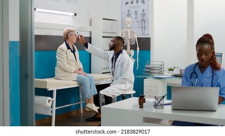 Male Physician Using Thermometer To Measure Temperature On Elderly Woman At Medical Consultation In Office. Doctor Doing Fever Measurement With Tool On Senior Patient At Cabinet.