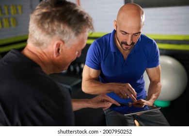 Male physio therapist and wellness personal trainer having a consultation with elderly senior man on strength and rehabilitation and assisted exercise routine in home gym looking at digital tablet