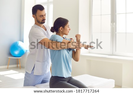 Male physiotherapist or chiropractor examining female patient's injured arm, stretching her muscles, helping her with medical exercise. Woman patient getting rehabilitation therapy in modern clinic