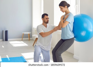 Male physiotherapist doctor helping female patient to do exercises with fitness ball in physiotherapy room. Woman crossed her arms and pressed the ball with her back to the wall to relieve the pain. - Powered by Shutterstock