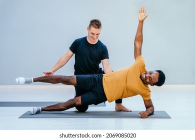 Male pilates trainer helps to african man, individual training participant, do right perform correctly. Extended different pose, class in sport club studio led by experienced man instructor concept.